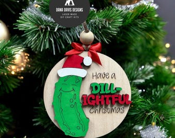 May include: Wooden Christmas ornament featuring a smiling green pickle wearing a Santa hat. The ornament reads "Have a Dill-ightful Christmas" in black and red lettering. A red bow and wooden bead are attached for hanging.