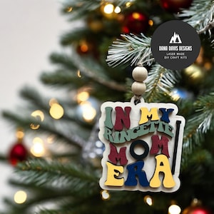 May include: A wooden Christmas tree ornament with the text "IN MY RINGETTE MOM ERA" in various colors. The ornament is hanging on a Christmas tree with colorful ornaments and lights. The ornament is made by Dana Davis Designs.