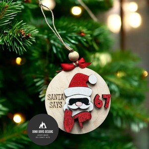 May include: Wooden Christmas bauble with a Santa Claus design. The bauble features a red Santa hat, sunglasses, and the text "SANTA SAYS 67". It is suspended from a string with a red bow and wooden bead. The bauble is set against a blurred Christmas tree background.