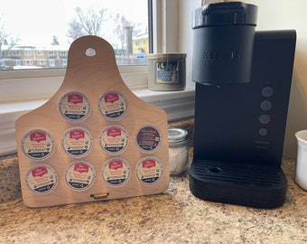 May include: A wooden coffee pod holder with a cut-out design in the shape of a deer's head. The holder has 10 slots for coffee pods and is filled with various Tim Hortons coffee pods. A black Keurig coffee maker is in the background.