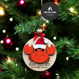 May include: A wooden Christmas ornament featuring a crab wearing a Santa hat. The crab is red, with a smiling face and large claws. The ornament has the text "MERRY CHRISTMAS YOU CRABBY BITCH!!" and is hanging from a Christmas tree.
