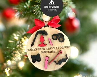 May include: A wooden Christmas ornament with a red ribbon and the text "Definatly on the naughty list this year SORRY Santa....". The ornament is decorated with various black and pink cutouts of suggestive items.