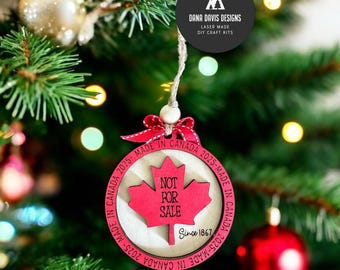 Pode incluir: Um enfeite de Natal de madeira vermelho e branco com um design de folha de bordo e o texto "Not For Sale Since 1867".