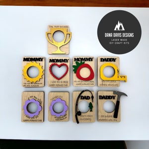 May include: A set of eight laser-cut wooden craft kits for creating personalized cards. Each card features a different design with a cutout for a photo or other embellishment. The designs include a sun, heart, strawberry, tape measure, flower, fishing rod, hammer, and a trophy. The cards are made of natural wood and have a rustic, handmade look.