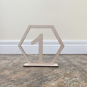 Puede incluir: Número de mesa de madera con un marco hexagonal y el número 1 recortado en el centro. El número de mesa está en un suelo de baldosas marrones.