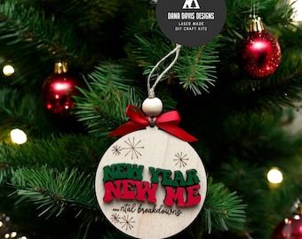 Puede incluir: Un adorno navideño de madera con el texto "Año Nuevo Yo Nuevo...desgastes mentales" en letras verdes y rojas. El adorno está colgado de un árbol de Navidad verde.