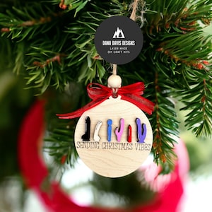 May include: A wooden Christmas ornament shaped like a circle with a red ribbon. The ornament has a white background with the text "Sending Christmas Vibes" and a row of colorful figures.