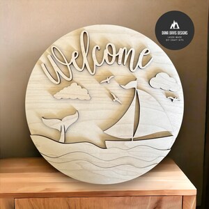 May include: A wooden round sign with the word "welcome" in a cursive font. The sign features a sailboat, a whale tail, and clouds, all cut out of the wood. The sign is perfect for a nautical themed home decor.