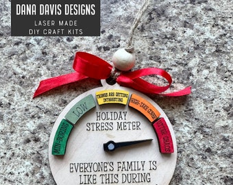 May include: A wooden holiday stress meter ornament with a red ribbon and a needle pointing to "utter chaos". The ornament is round and has a white background with black text that reads "Holiday Stress Meter" and "Everyone's family is like this during the holidays right?"