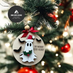May include: A wooden Christmas ornament with a ghost wearing a Santa hat and the text "BE MERRY STAY SCARY". The ornament has black bat cutouts and a red bow. The ornament is hanging on a Christmas tree with red ornaments.
