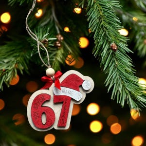 May include: Wooden Christmas tree decoration with a Santa hat design. The ornament features the number "67" in red, the word "NAME" on the hat, and the year "2023". It hangs from a string with a wooden bead and a red bow.
