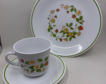 May include: A white ceramic cup, saucer, and plate set featuring a green rim and a pattern of red strawberries, green leaves, and yellow and white flowers. The plate has a central wreath design, while the cup and saucer feature matching motifs.