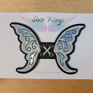 May include: A pair of iridescent silver butterfly shoe wings with black trim and a lace-up closure. The wings are attached to a black leather strap with eyelets. The text "Shoe Wings" and "TheCelestialGeek.com" are visible on the white background.