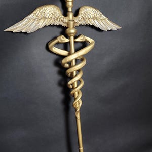Puede incluir: Un bastón de caduceo dorado con dos serpientes entrelazadas que se envuelven alrededor de una vara central, rematado con un par de alas detalladas y una pequeña esfera redonda de color marrón rojizo en la punta, sobre un fondo oscuro.