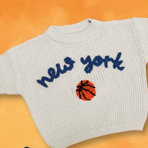 May include: Two children's sweaters, one cream and one navy blue, each with "new york" embroidered in contrasting colors. A basketball is embroidered below the text. The image includes the Statue of Liberty and the One World Trade Center.
