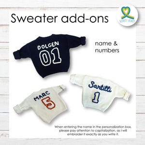 May include: Three white and navy blue sweaters with embroidered names and numbers. The sweaters are on a white wood background. The text on the sweaters reads "Dolgen 01", "Marc 5", and "Santilli 1".