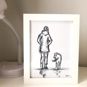 Op de afbeelding: Een zwart-wit schets in een witte lijst van een persoon met een hond. De persoon draagt een jas met capuchon en een broek. De hond kijkt naar de grond. De tekening is in een witte lijst.