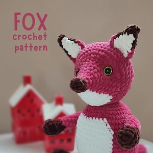 Puede incluir: Un patrón de crochet para un zorro rosa con detalles blancos y marrones. El patrón está disponible para descargar en PDF. El zorro está sentado sobre una superficie blanca.