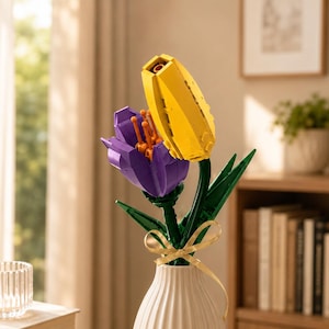 May include: A white, ribbed ceramic vase holds a vibrant arrangement of Lego flowers. The flowers include a yellow tulip and a purple flower with orange accents, complemented by green stems and leaves. A gold ribbon adorns the vase.