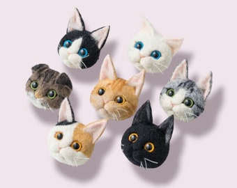 Könnte beinhalten: Eine Sammlung von sieben Filz-Katzenkopf-Ornamenten in verschiedenen Farben und Mustern. Die Katzen haben detaillierte Merkmale, einschließlich verschiedener Augenfarben und -zeichnungen. Die Katzen sind auf einem hellrosa Hintergrund angeordnet.