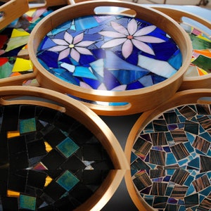 May include: A collection of round wooden serving trays with mosaic tile designs. The trays feature various mosaic patterns, including floral and abstract designs in shades of blue, black, and brown. The wooden rims have built-in handles.