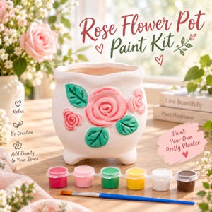 May include: A white ceramic flower pot with raised rose and leaf designs, accompanied by a strip of six small paint pots in red, pink, green, yellow, white, and brown, plus a blue-handled paintbrush. Text reads Rose Flower Paint Kit.