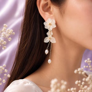 May include: A long drop earring featuring a gold-toned filigree flower at the top, a smaller floral accent below, and two delicate gold chains ending in small, white, oval-shaped pearls that hang at different lengths from the flower base.