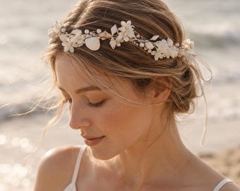 May include: A floral hair crown featuring white blossoms, small seashells, and round white pearls arranged on a thin metallic wire base. The accessory includes delicate white ribbon ties at the ends, creating a coastal-inspired headpiece.