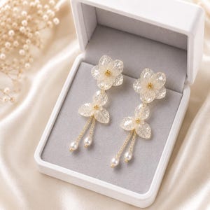 May include: A pair of drop earrings featuring translucent, skeletonised leaf-shaped petals arranged in flower patterns. Each earring includes a small gold-toned centre, delicate gold chains, and two dangling white freshwater pearls.