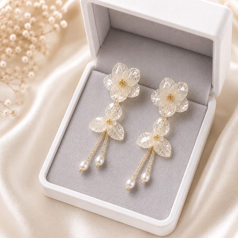 May include: A pair of drop earrings featuring translucent, skeletonised leaf-shaped petals arranged in flower patterns. Each earring includes a small gold-toned centre, delicate gold chains, and two dangling white freshwater pearls.