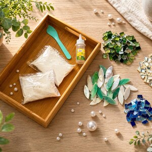 May include: A wooden tray holds a teal spoon, glue bottle, and two bags of white material. Scattered around are glass mosaic tiles in green, blue, and iridescent colors, along with faux pearls. The scene is set on a light wood surface.
