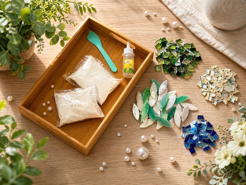 May include: A wooden tray holds a teal spoon, glue bottle, and two bags of white material. Scattered around are glass mosaic tiles in green, blue, and iridescent colors, along with faux pearls. The scene is set on a light wood surface.