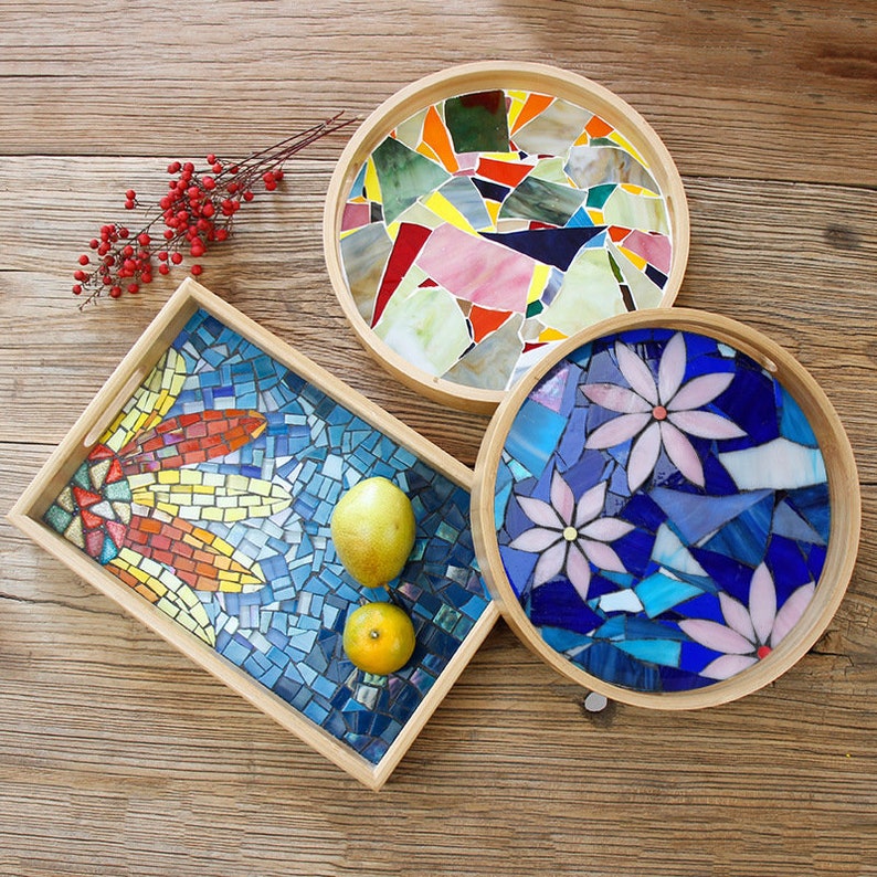 May include: Three decorative mosaic trays with wooden frames. One rectangular tray features a dragonfly design, while two round trays showcase abstract and floral patterns. The mosaic tiles are in various colors, including blue, orange, and pink.