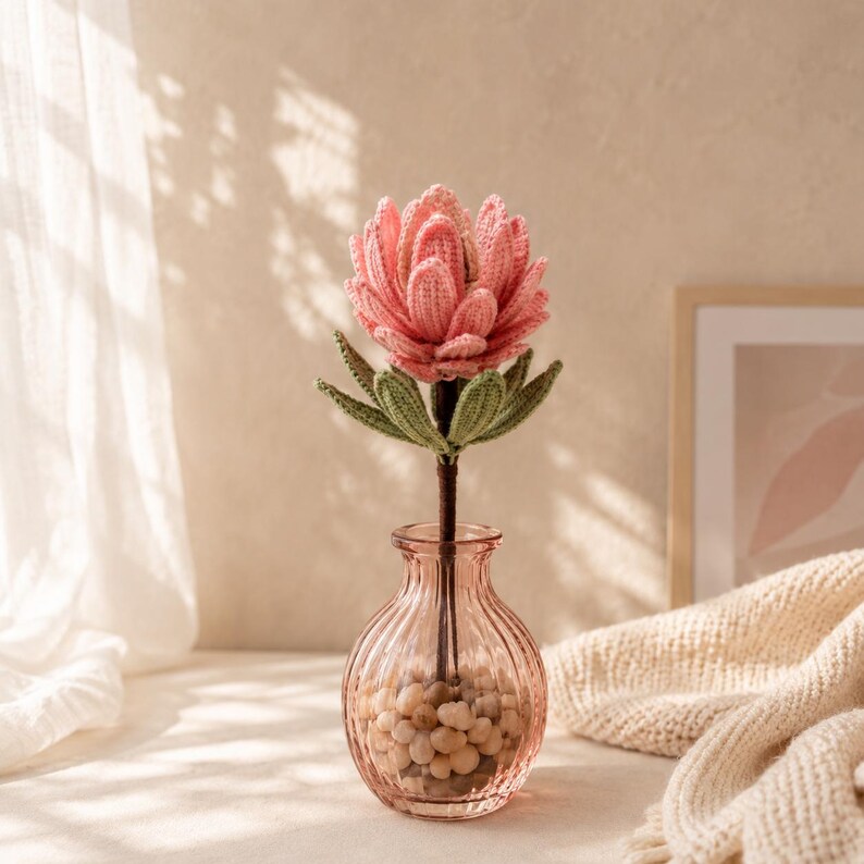 May include: A crocheted pink protea flower with green leaves on a dark brown stem, placed inside a clear, ribbed glass vase filled with small, light-colored pebbles. The flower features intricate yarn textures and layered petals.