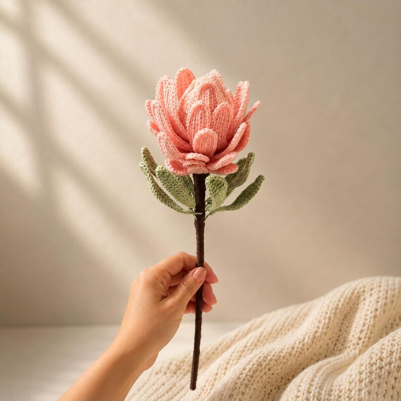 May include: A handcrafted crochet protea flower featuring layered, soft pink petals and textured sage green leaves attached to a dark brown stem. The intricate yarn work creates a realistic floral structure with a dense, rounded bloom and pointed foliage.