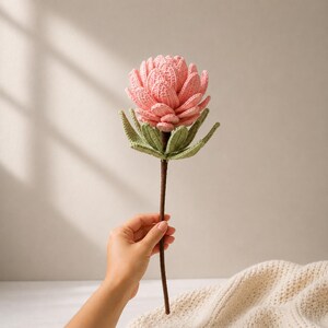 May include: A handcrafted crochet flower featuring a layered, light pink bloom with textured petals. The flower has a set of light green leaves at the base of the bloom and is attached to a long, thin, dark brown stem, creating a botanical display.