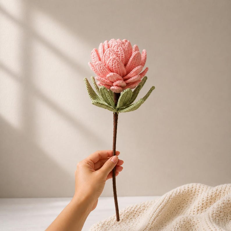 May include: A handcrafted crochet flower featuring a layered, light pink bloom with textured petals. The flower has a set of light green leaves at the base of the bloom and is attached to a long, thin, dark brown stem, creating a botanical display.