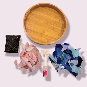 May include: A craft kit with a round bamboo tray, a black bag, a pink floral mosaic, a pile of blue mosaic tiles, glue, and small round pieces. The kit is designed for creating mosaic art projects.