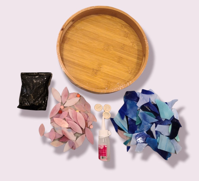 May include: A craft kit with a round bamboo tray, a black bag, a pink floral mosaic, a pile of blue mosaic tiles, glue, and small round pieces. The kit is designed for creating mosaic art projects.