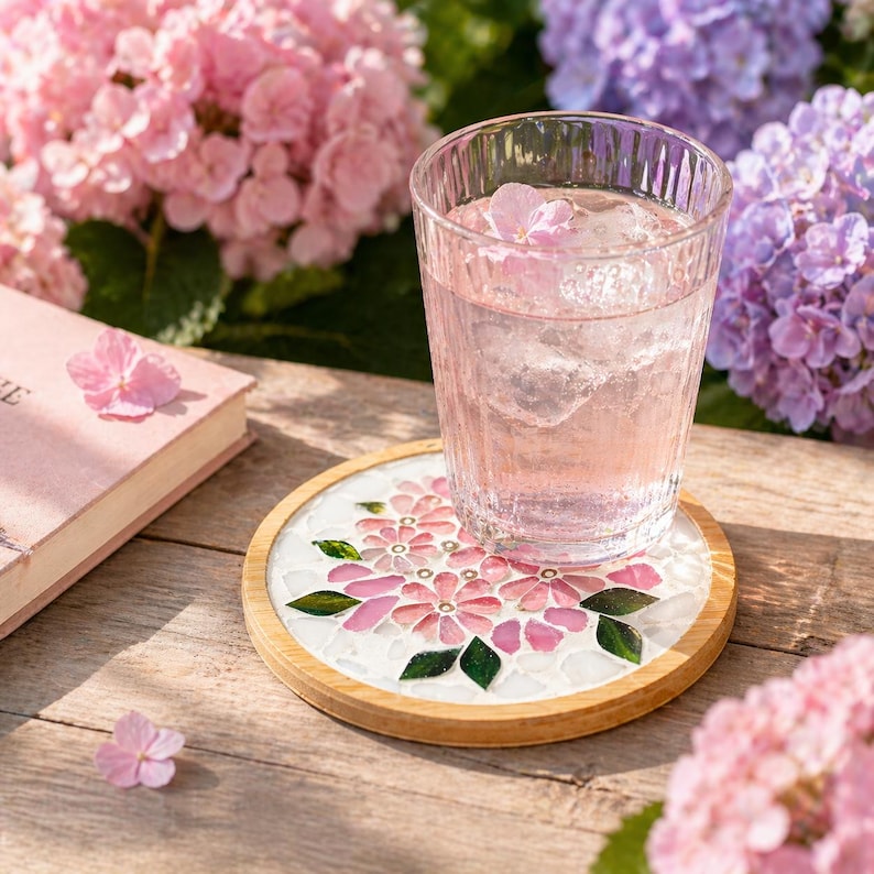 May include: A glass of pink liquid with ice and a flower sits on a floral mosaic coaster with a wooden rim. The coaster features pink flowers and green leaves on a white background. Pink flowers and a book are in the background.