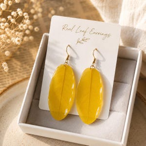 May include: A pair of yellow leaf-shaped earrings with gold-toned metal hooks, displayed on a white rectangular card labeled Real Leaf Earrings. The earrings feature a smooth, translucent finish showing natural leaf veins, presented in a box.