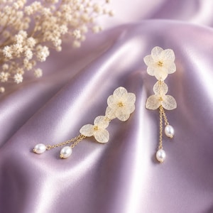 May include: A pair of gold-toned drop earrings featuring delicate, translucent, cream-coloured flower petals with fine veining. Each earring is accented with small, lustrous white pearls suspended from thin gold chains, creating a layered floral design.