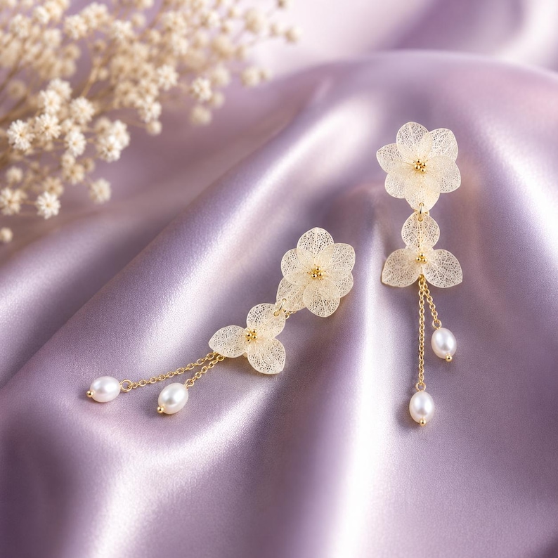 May include: A pair of gold-toned drop earrings featuring delicate, translucent, cream-coloured flower petals with fine veining. Each earring is accented with small, lustrous white pearls suspended from thin gold chains, creating a layered floral design.