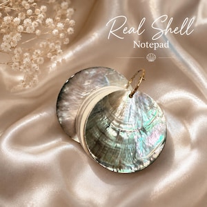 May include: A circular notepad featuring two iridescent, polished seashell covers with a natural pearlescent finish. The pages are bound together by a gold-toned metal ring. The text Real Shell Notepad is printed in white script above the object.