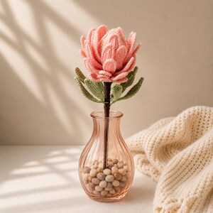 May include: A crocheted pink protea flower with green leaves on a dark brown stem, placed inside a clear, ribbed glass vase filled with small, light-colored pebbles. The flower features layered, textured petals crafted from yarn.