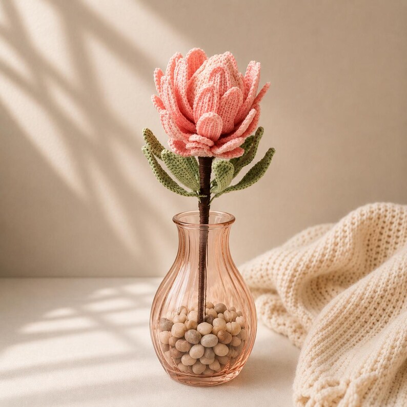 May include: A crocheted pink protea flower with green leaves on a dark brown stem, placed inside a clear, ribbed glass vase filled with small, light-colored pebbles. The flower features layered, textured petals crafted from yarn.