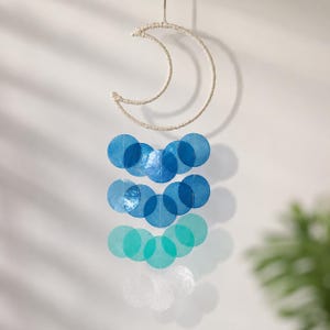 Puede incluir: Un adorno colgante de pared con forma de media luna envuelta en hilo natural. Debajo, una cascada de discos circulares translúcidos en tonos azules, turquesas y blancos crea un efecto degradado. El diseño está suspendido por un lazo de hilo a juego.