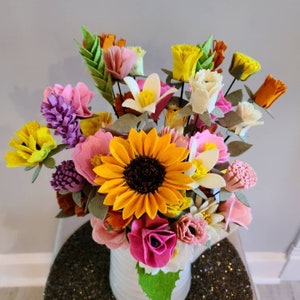 May include: A vibrant bouquet of felt flowers in a white vase. The arrangement includes a large yellow sunflower, pink roses, and various other colorful blooms. The flowers are arranged in a decorative vase, creating a cheerful display.