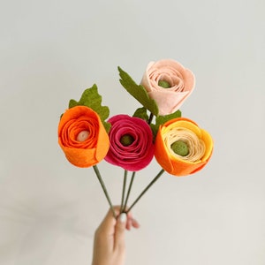 Pu&ograve; includere: Un bouquet di fiori in feltro fatti a mano nei toni dell'arancione, del rosa e del rosso. I fiori hanno steli e foglie verdi. Il bouquet &egrave; tenuto su uno sfondo bianco.