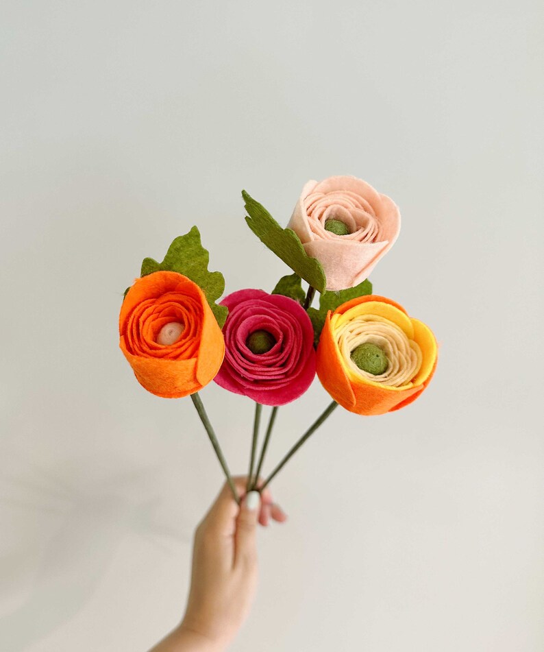 Pu&ograve; includere: Un bouquet di fiori in feltro fatti a mano nei toni dell'arancione, del rosa e del rosso. I fiori hanno steli e foglie verdi. Il bouquet &egrave; tenuto su uno sfondo bianco.